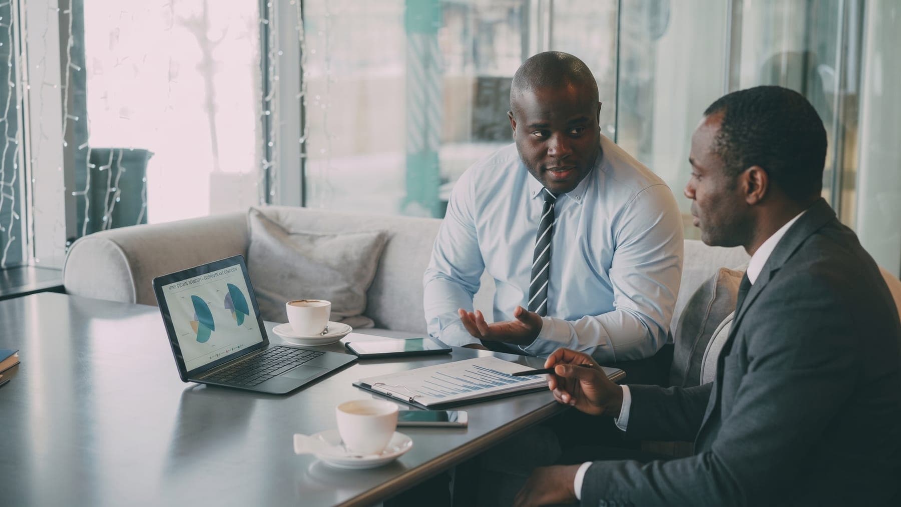 Two people across a table with a laptop of charts and a printed report between them. The deal being argued, not the software.